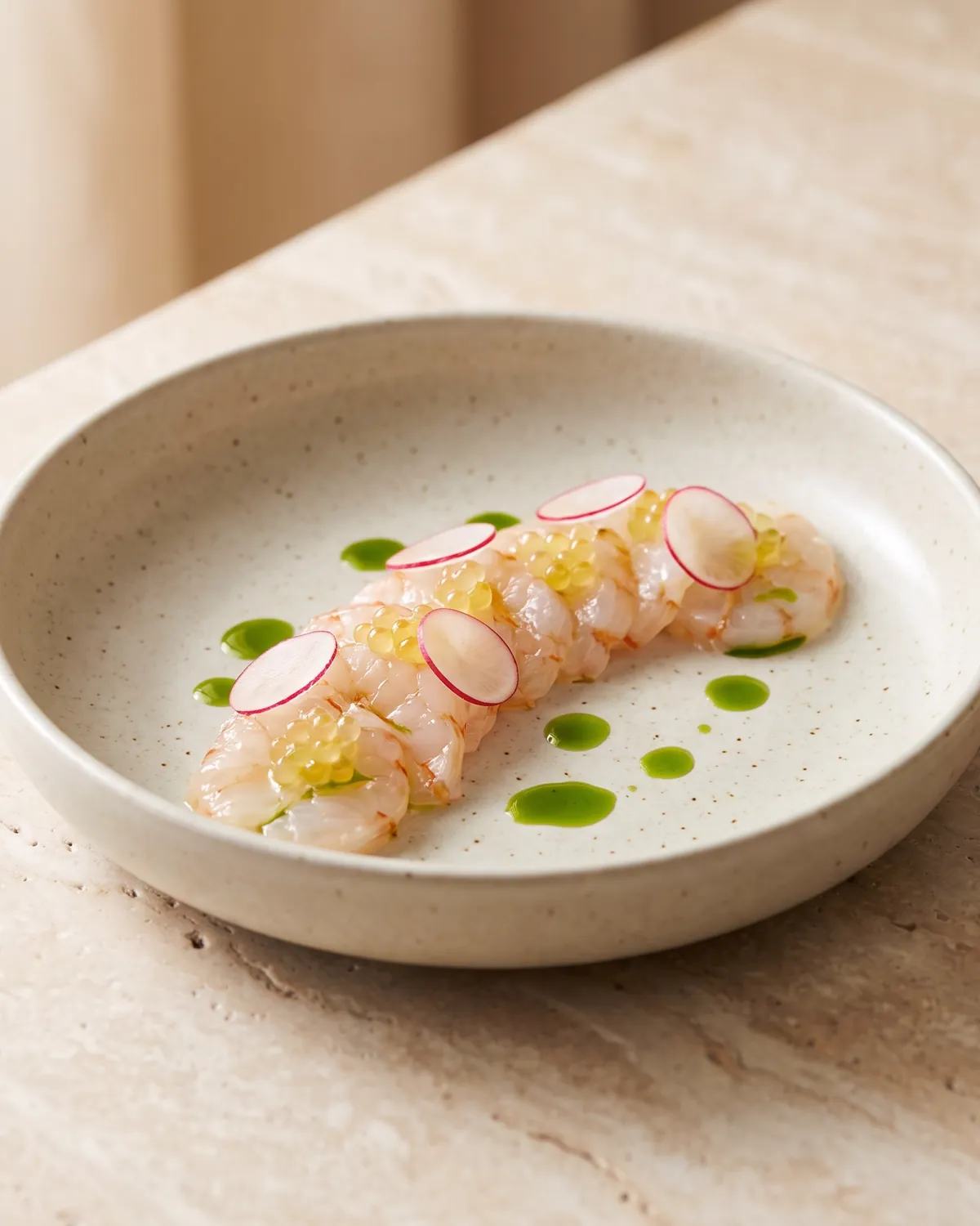 Slices of raw langoustine with shaved radish and drops of green herb oil in a wide ceramic bowl.